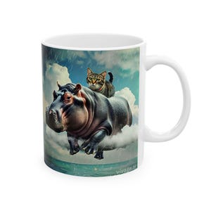 May include: White ceramic mug with a fantasy illustration of a hippo and a cat in a cloudy sky above the sea. The hippo is grey, and the cat is brown and tan. The mug has a white handle.