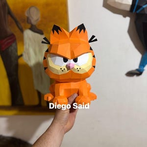 K&ouml;nnte beinhalten: Eine orangefarbene Garfield-Figur aus Papier mit schwarzen Streifen, Ohren und einer rosa Nase. Die Figur hat einen w&uuml;tenden Gesichtsausdruck und der Text "Diego Said" ist auf der Basis sichtbar. Es ist eine 3D-Papierkunst.