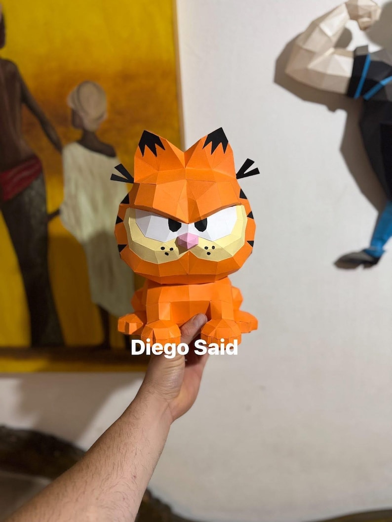 K&ouml;nnte beinhalten: Eine orangefarbene Garfield-Figur aus Papier mit schwarzen Streifen, Ohren und einer rosa Nase. Die Figur hat einen w&uuml;tenden Gesichtsausdruck und der Text "Diego Said" ist auf der Basis sichtbar. Es ist eine 3D-Papierkunst.