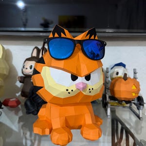 K&ouml;nnte beinhalten: Eine geometrische Papierkunst-Skulptur von Garfield in Orange, die eine schwarze Sonnenbrille mit blauen Gl&auml;sern tr&auml;gt. Die Skulptur hat schwarze Streifen, eine rosa Nase und drei schwarze Punkte auf jeder Wange. Andere Papierkunstfiguren sind im Hintergrund.