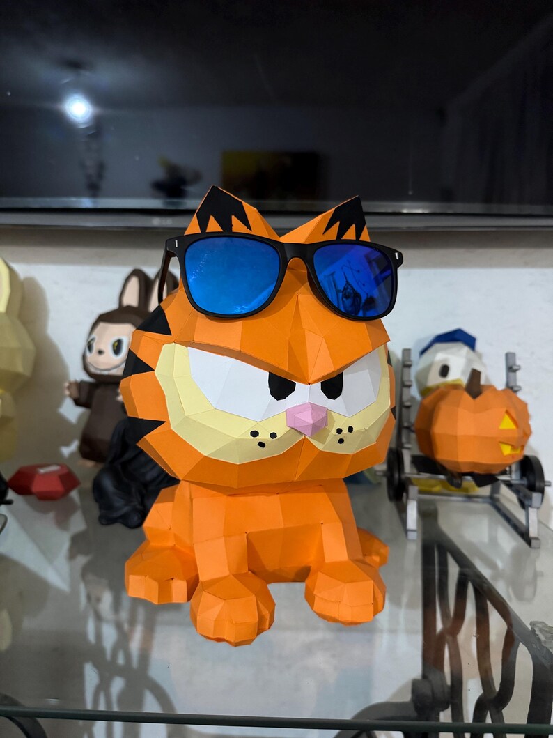 K&ouml;nnte beinhalten: Eine geometrische Papierkunst-Skulptur von Garfield in Orange, die eine schwarze Sonnenbrille mit blauen Gl&auml;sern tr&auml;gt. Die Skulptur hat schwarze Streifen, eine rosa Nase und drei schwarze Punkte auf jeder Wange. Andere Papierkunstfiguren sind im Hintergrund.