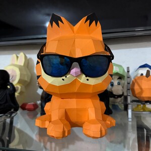 K&ouml;nnte beinhalten: Eine orangefarbene Papierfigur von Garfield der Katze mit schwarzer Sonnenbrille. Die Figur hat schwarze Markierungen auf den Ohren und eine rosa Nase. Weitere Zeichentrickfiguren sind im Hintergrund zu sehen.