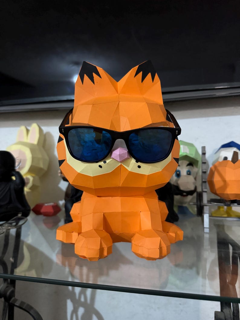 K&ouml;nnte beinhalten: Eine orangefarbene Papierfigur von Garfield der Katze mit schwarzer Sonnenbrille. Die Figur hat schwarze Markierungen auf den Ohren und eine rosa Nase. Weitere Zeichentrickfiguren sind im Hintergrund zu sehen.