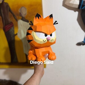 K&ouml;nnte beinhalten: Eine orangefarbene Garfield-Figur aus Papier mit schwarzen Streifen und einer rosa Nase. Die Figur wird von einer Hand gehalten. Der Text "Diego Said" ist auf dem Bild zu sehen.