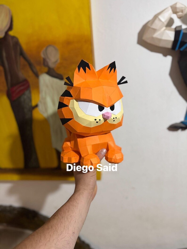 K&ouml;nnte beinhalten: Eine orangefarbene Garfield-Figur aus Papier mit schwarzen Streifen und einer rosa Nase. Die Figur wird von einer Hand gehalten. Der Text "Diego Said" ist auf dem Bild zu sehen.