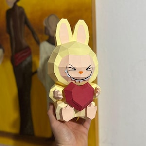Puede incluir: Una figura de conejito de papel amarillo con un corazón rojo. El conejito tiene una cara sonriente con detalles negros y sostiene un corazón rojo. La figura está hecha de papel plegado y tiene un diseño geométrico.
