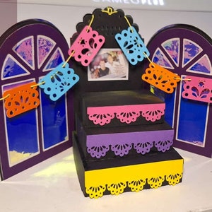 May include: A three-tiered black display stand with scalloped pink, purple, and yellow paper trim. Behind the stand are two purple arched window frames with blue inserts, connected by a string of colorful, intricate paper cut-out flags.