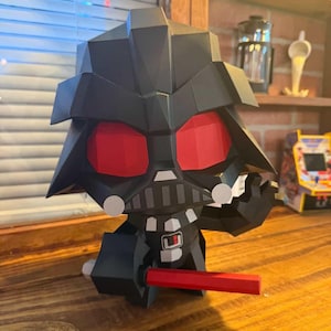 May include: A black papercraft figure of Darth Vader with red eyes and a red lightsaber. The figure is in a seated position with a detailed helmet and chest plate. The figure is on a wooden surface.