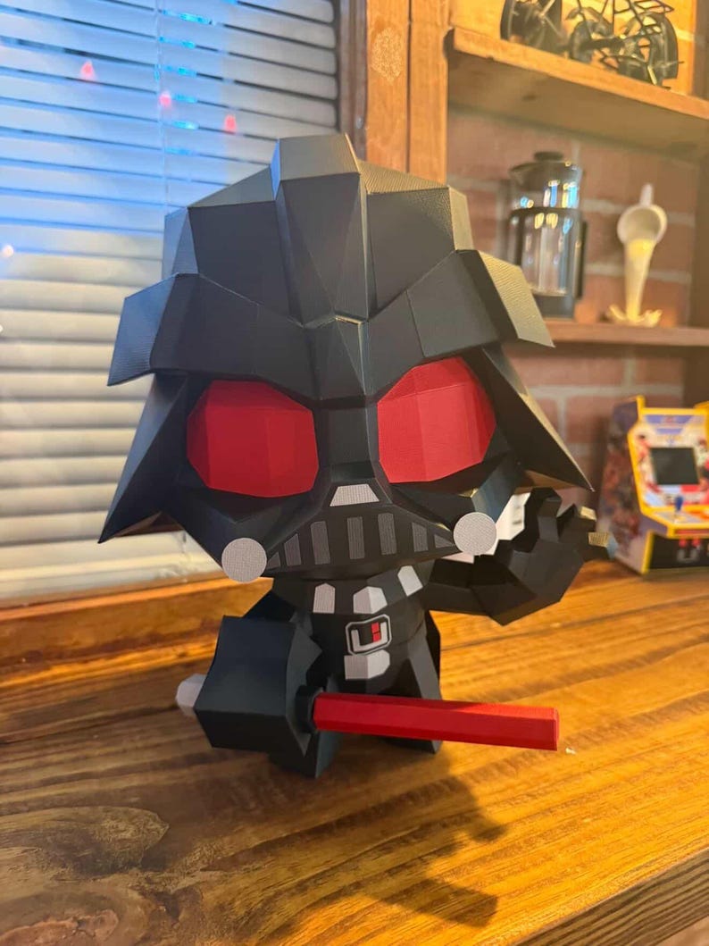May include: A black papercraft figure of Darth Vader with red eyes and a red lightsaber. The figure is in a seated position with a detailed helmet and chest plate. The figure is on a wooden surface.
