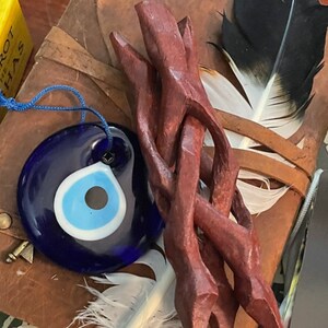 Op de afbeelding: Een blauwe glazen boze oog charme met een wit en lichtblauw oogontwerp, hangend aan een blauw koord. De charme is op een bruin lederen dagboek met een houten stok met een gevlochten ontwerp.