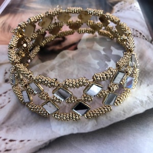 Puede incluir: Pulsera dorada con tres filas. La pulsera presenta pequeñas cuentas redondas y detalles de espejo en forma de diamante. La joya se exhibe sobre una superficie suave y texturizada, lo que sugiere un enfoque en los accesorios de moda.