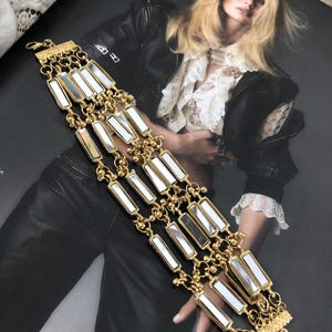 May include: Gold-toned bracelet featuring rectangular mirrored accents. The bracelet has multiple strands of delicate chain and rectangular mirrored pieces. The bracelet is displayed on a magazine page with a fashion model.