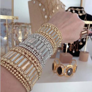 Puede incluir: Una colección de pulseras doradas y plateadas, con diseños de cuentas y rectangulares, apiladas en una muñeca. Otras joyas de oro, incluyendo collares y una pulsera con piedras oscuras, se exhiben en el fondo.