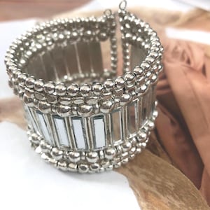 Puede incluir: Pulsera de puño plateada con un diseño en capas. La pulsera presenta filas de pequeñas cuentas redondas y detalles rectangulares espejados. El brazalete es ancho y tiene un diseño flexible.