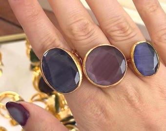 May include: Three gold-toned rings with large, faceted gemstones in shades of purple and blue. The rings are displayed on a hand with polished nails. Other rings are visible in the background, suggesting a jewelry collection.