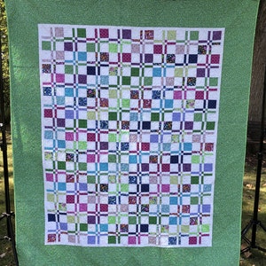 May include: A green and white quilt with a patchwork design. The quilt is made up of squares of different fabrics, each with a different pattern. The quilt is displayed on a stand in an outdoor setting.