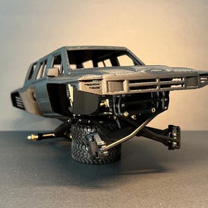 May include: A dark gray 3D printed model of a truck body featuring an open cabin, a front grille, and side vents. The chassis is exposed, showing black suspension components and metal hardware, while the vehicle rests on two stacked black tires.