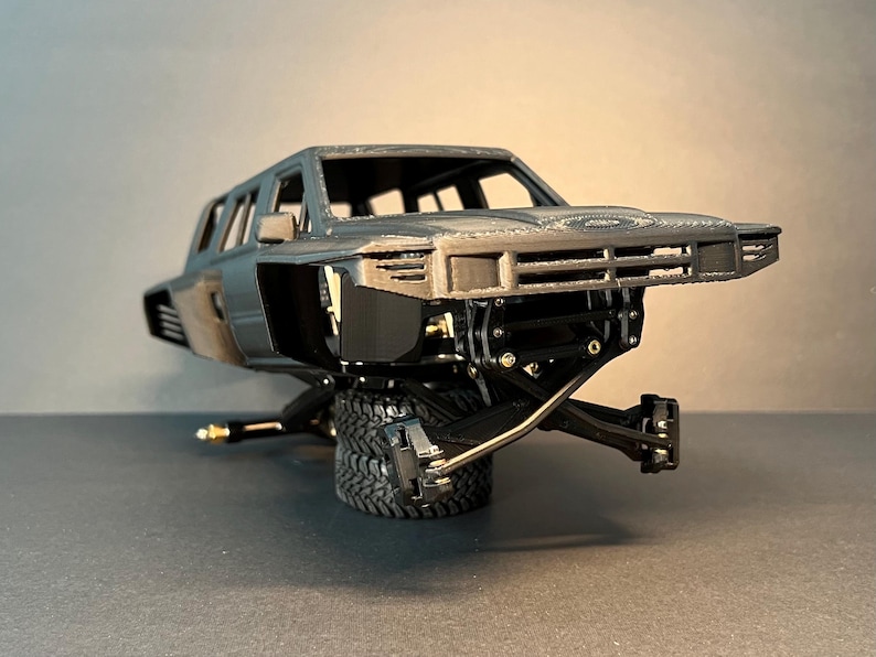 May include: A dark gray 3D printed model of a truck body featuring an open cabin, a front grille, and side vents. The chassis is exposed, showing black suspension components and metal hardware, while the vehicle rests on two stacked black tires.