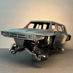May include: A dark gray 3D printed model car body featuring a wide-body kit, an open cabin, and exposed front suspension components. The vehicle chassis is mounted on a single black off-road tire, showcasing detailed mechanical parts.