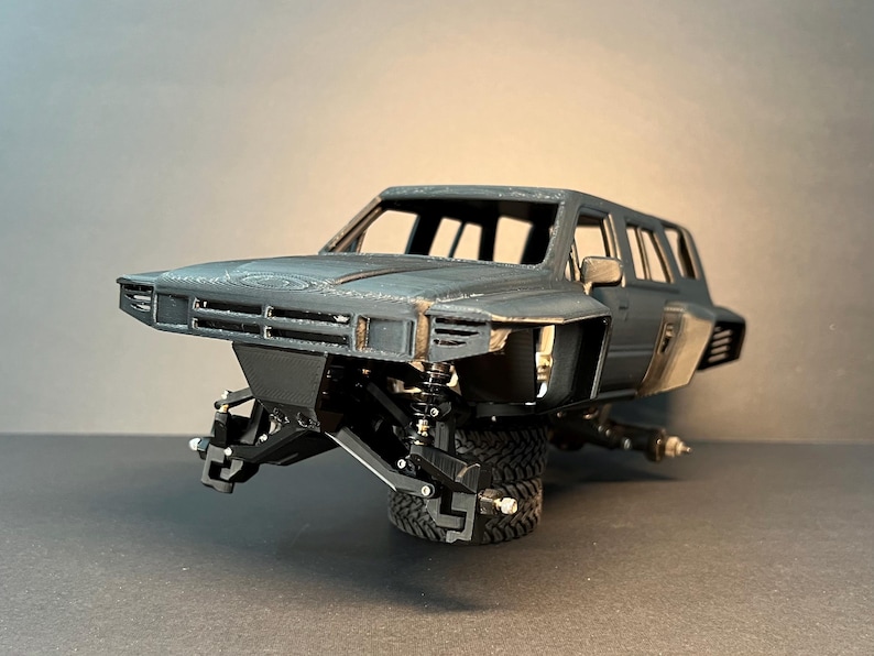 May include: A dark gray 3D printed model car body featuring a wide-body kit, an open cabin, and exposed front suspension components. The vehicle chassis is mounted on a single black off-road tire, showcasing detailed mechanical parts.