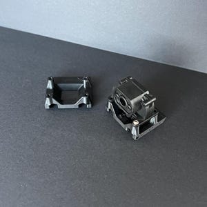 Könnte beinhalten: Zwei schwarze mechanische Kunststoffkomponenten werden vor einem dunklen Hintergrund gezeigt. Eine Komponente ist eine quadratische Basis mit einer vertieften Mitte und vier Schraubenlöchern. Die andere Komponente hat ein zylindrisches Element und ist auf einer ähnlichen Basis montiert.
