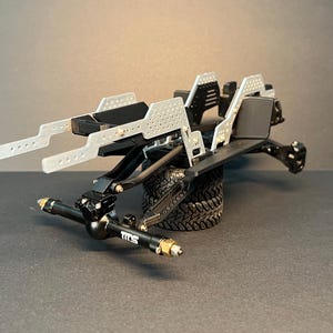 May include: A black and silver radio-controlled vehicle chassis featuring a perforated metal frame, black suspension components, and a front axle marked with the text TITS. The assembly is supported by a single black rubber tire with a deep tread.