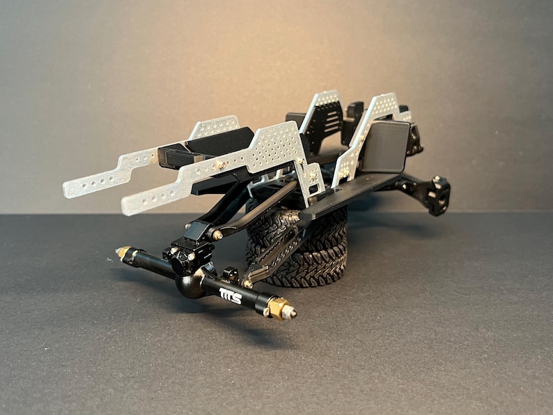 May include: A black and silver radio-controlled vehicle chassis featuring a perforated metal frame, black suspension components, and a front axle marked with the text TITS. The assembly is supported by a single black rubber tire with a deep tread.