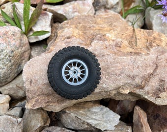 Könnte beinhalten: Ein schwarzer und grauer Geländereifen mit einem hellgrauen Rad, der auf einer Ansammlung von Steinen liegt. Der Reifen hat ein detailliertes Profil und die Wörter "All Terrain M/T" und "Gravel Blaster" sind sichtbar.