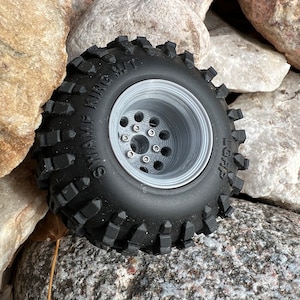 May include: A black off-road tire with a silver rim. The tire has a deep tread pattern and the words "SWAMP KING MT" are printed on the sidewall. The rim has multiple holes and is designed for a vehicle.