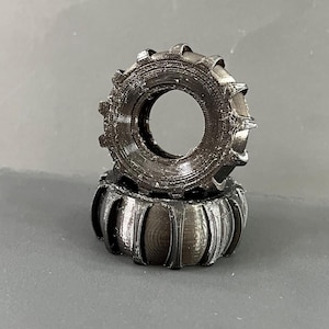 Könnte beinhalten: Zwei dunkelgraue, zahnradförmige Objekte sind übereinander gestapelt. Das obere Zahnrad hat ein großes Mittelloch und einen geriffelten Außenrand. Das untere Zahnrad hat ein ähnliches Design mit einer Reihe von gebogenen Rippen. Die Objekte scheinen aus einem strukturierten Material zu bestehen.