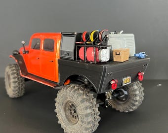 Op de afbeelding: Een miniatuur oranje en zwarte speelgoedvrachtwagen met grote, robuuste banden. De laadbak van de vrachtwagen is beladen met apparatuur, waaronder rode tanks, een bruine doos en een zilveren container. De vrachtwagen heeft een kentekenplaat met "FOR FUN".