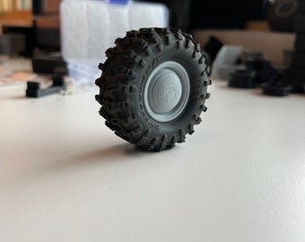 Op de afbeelding: Een grijze terreinwagenband met een gedetailleerd profiel. De band is gemonteerd op een grijze velg. De band is ontworpen voor ruig terrein en is waarschijnlijk voor een modelvoertuig.