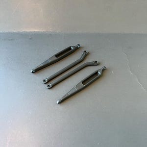 以下が含まれることがあります： 4つの黒い細長い金属製の物体が灰色の表面に配置されています。2つは長方形の開口部を持つ直線状で、2つは円形の開口部を持つ曲線状です。これらはコンポーネントまたは部品のようです。