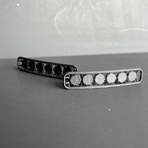 Könnte beinhalten: Zwei gebogene, 3D-gedruckte Lichtleisten in Schwarz und Grau. Jede Leiste hat sieben kreisförmige Lichtelemente. Die Leisten liegen auf einer grauen Oberfläche.