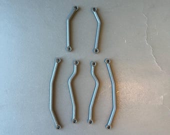 Op de afbeelding: Een verzameling van acht kleine, donkergrijze metalen staven. De staven hebben verschillende vormen, waaronder rechte, gebogen en gebogen ontwerpen. Elke staaf heeft aan elk uiteinde een klein gaatje. De staven zijn op een grijze ondergrond geplaatst.
