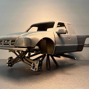 May include: A gray 3D-printed model of a pickup truck with a lifted suspension. The truck has a detailed front end, side mirrors, and a bed. The suspension is black and features intricate mechanical details.