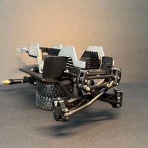 May include: A black and silver model vehicle chassis featuring a complex suspension system, exposed mechanical linkages, and a textured frame. The structure includes two stacked black rubber tires and silver perforated panels on the upper section.