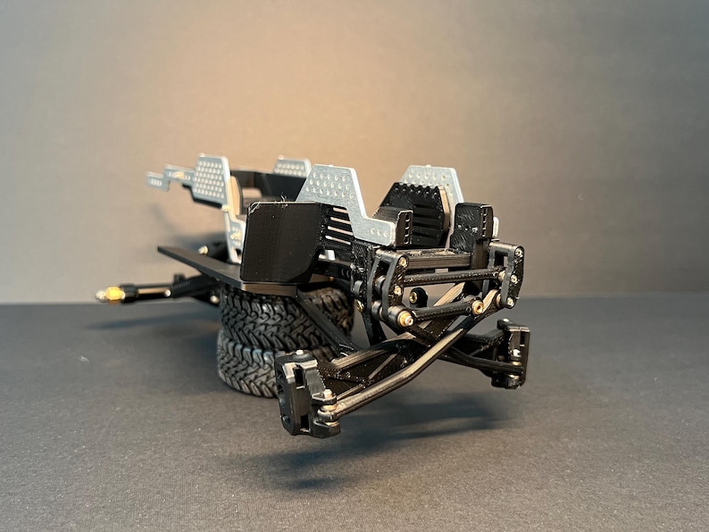 May include: A black and silver model vehicle chassis featuring a complex suspension system, exposed mechanical linkages, and a textured frame. The structure includes two stacked black rubber tires and silver perforated panels on the upper section.