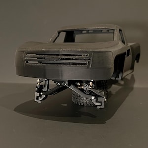Könnte beinhalten: Ein schwarzes, unfertiges Modell-LKW-Chassis mit sichtbarer Aufhängung und Rädern. Die Vorderseite des Trucks weist einen detaillierten Kühlergrill und Scheinwerfer auf. Das Modell ist vor einem dunklen Hintergrund platziert.