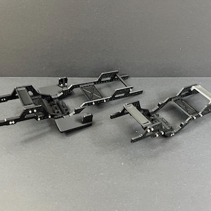 Puede incluir: Dos chasis de modelo de coche de metal negro. Los marcos tienen forma rectangular con múltiples agujeros y puntos de montaje. Los chasis están diseñados para un vehículo a pequeña escala, con elementos estructurales detallados y un acabado mate.
