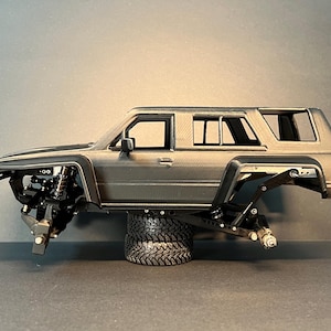 May include: A gray scale model of a four-door SUV body shell featuring a textured finish, black wheel arches, and an exposed chassis suspension system. The vehicle frame is supported by a stack of two black rubber tires, showcasing the mechanical components.