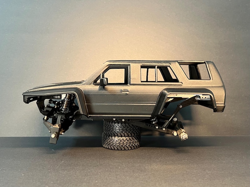 May include: A gray scale model of a four-door SUV body shell featuring a textured finish, black wheel arches, and an exposed chassis suspension system. The vehicle frame is supported by a stack of two black rubber tires, showcasing the mechanical components.