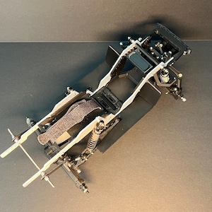 May include: A black and silver remote-controlled car chassis featuring a central frame, exposed suspension components, coil-over shock absorbers, and a textured battery strap. The structure includes front and rear axle mounts with metal linkages.