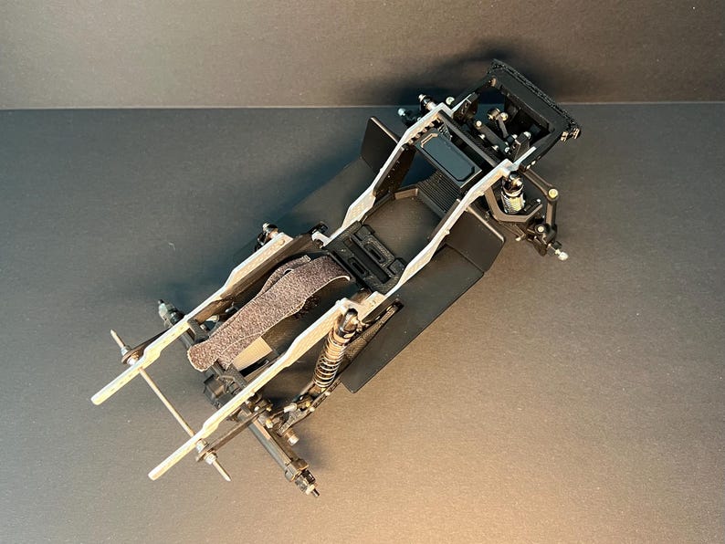 May include: A black and silver remote-controlled car chassis featuring a central frame, exposed suspension components, coil-over shock absorbers, and a textured battery strap. The structure includes front and rear axle mounts with metal linkages.