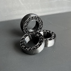 Könnte beinhalten: Drei schwarze, kreisförmige Objekte mit einem strukturierten, wabenartigen Muster am äußeren Rand. Die Objekte sind auf einer grauen Oberfläche gestapelt und angeordnet, was eine interessante Komposition ergibt. Die Objekte scheinen aus mattem Material zu bestehen.