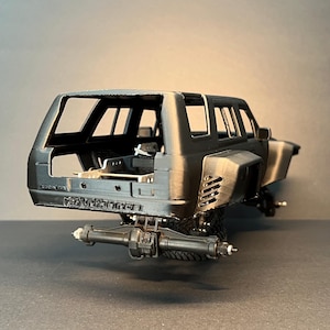 May include: A matte black scale model vehicle body featuring a wide-body design with rear fender vents, an open interior cabin, and a rear axle assembly. The rear bumper area displays the embossed text "STRYKER" in a bold, capitalized font.