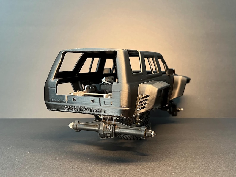 May include: A matte black scale model vehicle body featuring a wide-body design with rear fender vents, an open interior cabin, and a rear axle assembly. The rear bumper area displays the embossed text "STRYKER" in a bold, capitalized font.