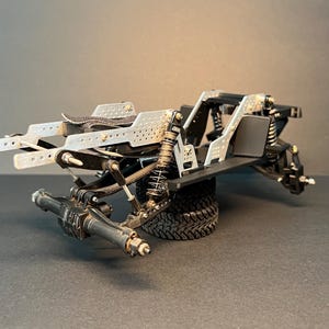 May include: A remote-controlled vehicle chassis featuring a silver perforated metal frame, black suspension components, coil-over shocks, and a black rear axle assembly. The structure is supported by a single black rubber tire with a deep tread pattern.