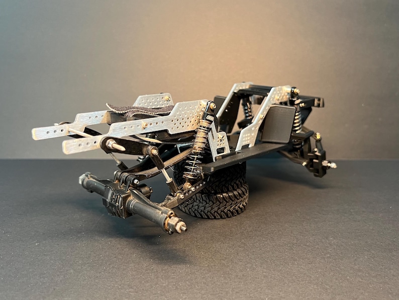May include: A remote-controlled vehicle chassis featuring a silver perforated metal frame, black suspension components, coil-over shocks, and a black rear axle assembly. The structure is supported by a single black rubber tire with a deep tread pattern.
