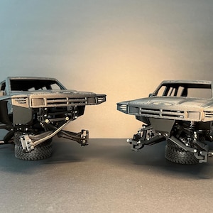 May include: Two dark gray 3D-printed model truck bodies featuring a detailed front grille, hood, and cabin structure. Each model is mounted on a single black tire, showcasing the exposed front suspension components, control arms, and steering knuckles.